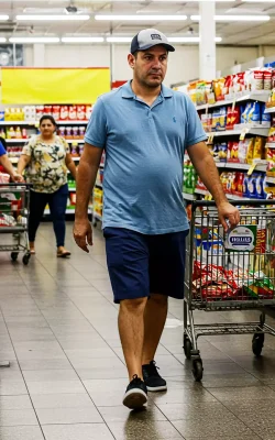 Homem andando em um supermercado com carrinho de compras, demonstrando desinteresse ou indecisão em um ambiente de prateleiras com produtos sem precificação aparente.