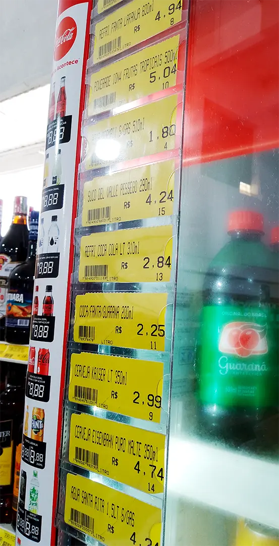 Porta-etiquetas verticais com preços destacados de bebidas em refrigerador de supermercado, com etiquetas amarelas bem visíveis.