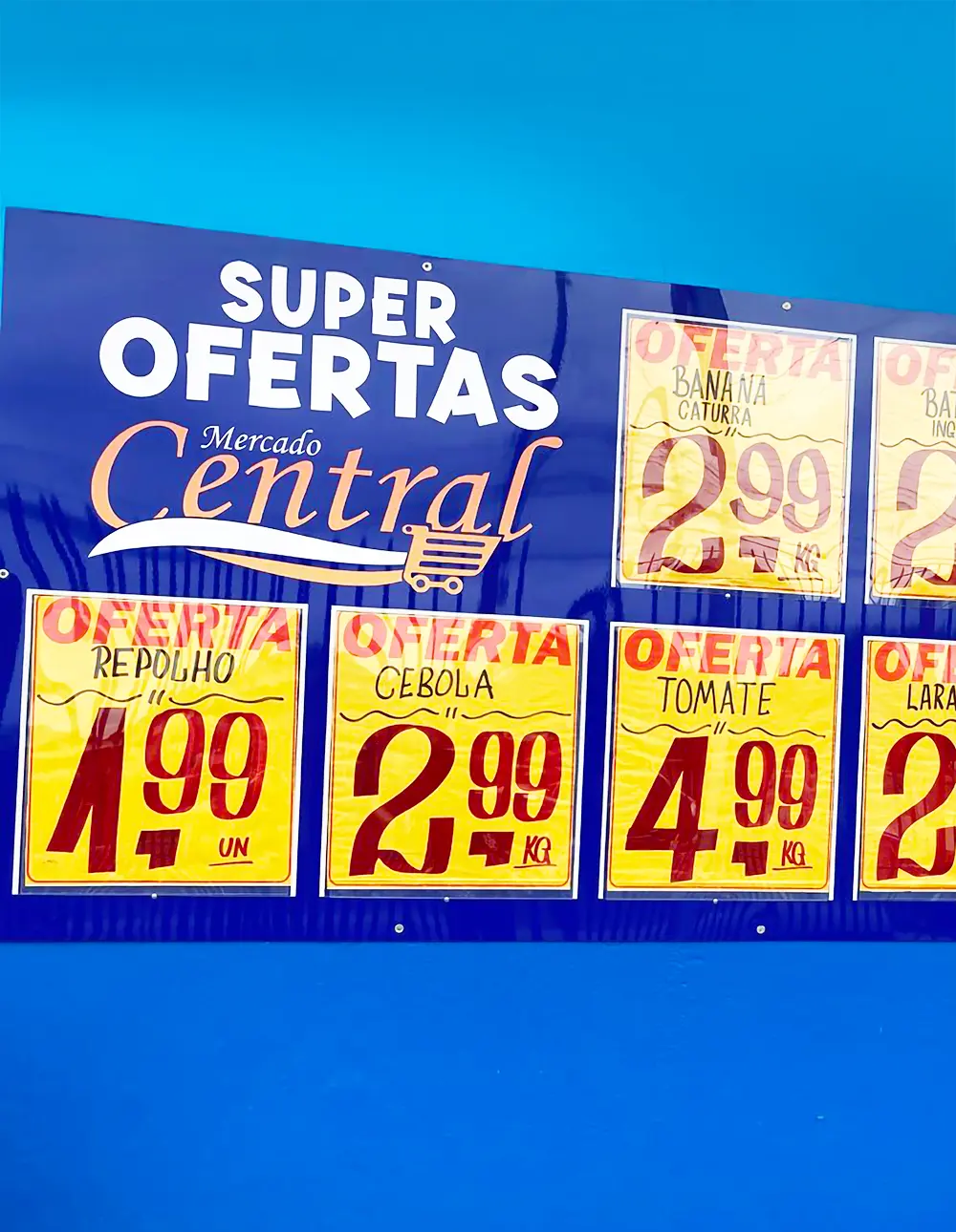 Painel com porta-cartazes exibindo ofertas de frutas, legumes e verduras em um supermercado, com fundo azul e destaque para os preços.