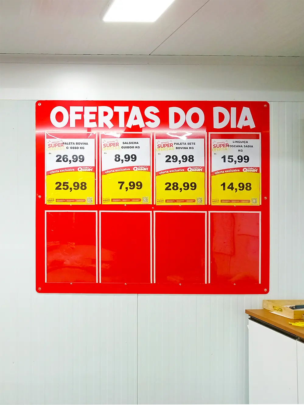 Painel de porta-cartazes vermelho com o título “Ofertas do Dia”, exibindo promoções de carnes e embutidos em um supermercado.