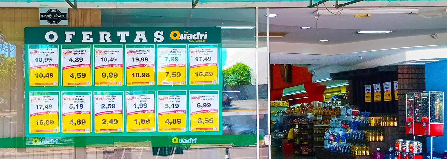 Painel com porta-cartazes exibindo ofertas na vitrine de entrada do Supermercado Quadri, com destaque para promoções de cervejas, arroz, feijão e açúcar.
