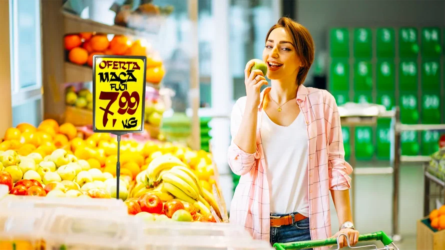 Cliente sorrindo ao lado de um cartaz de oferta de maçã Fuji por R$ 7,99 em um supermercado, ilustrando a importância da visibilidade das promoções para aumentar as vendas.