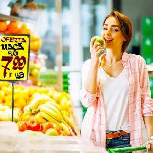 Cliente sorrindo ao lado de um cartaz de oferta de maçã Fuji por R$ 7,99 em um supermercado, ilustrando a importância da visibilidade das promoções para aumentar as vendas.