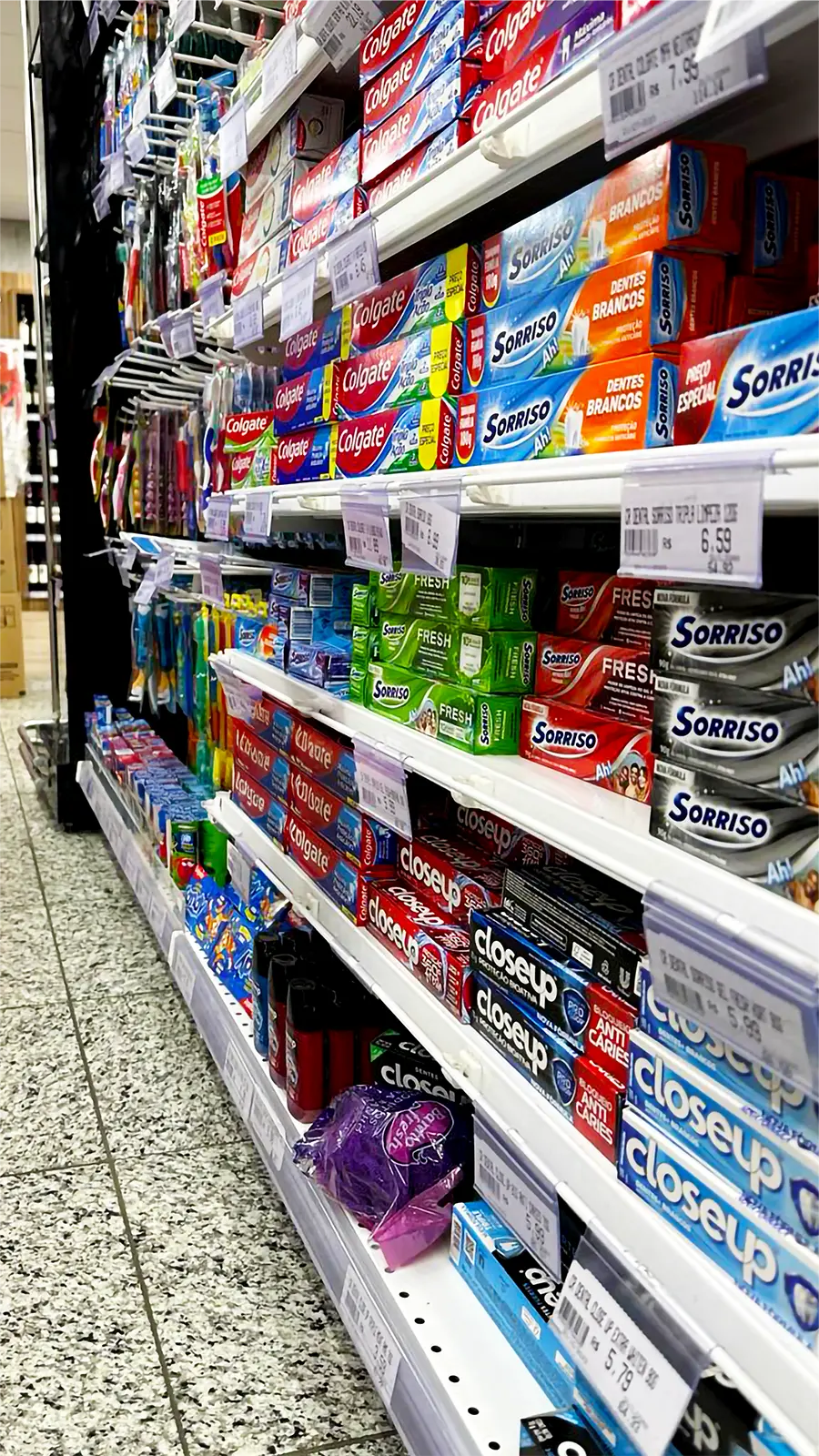 Prateleira de supermercado lotada de pastas de dente (Colgate, Sorriso, Closeup) com etiquetas eletrônicas de preço claramente visíveis.