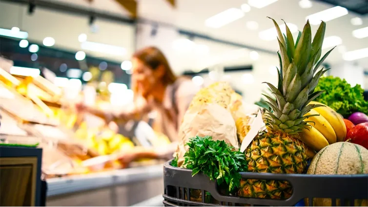 Carrinho cheio de frutas e verduras em supermercado com cliente ao fundo escolhendo produtos, ilustrando a comunicação visual para aumentar ticket médio no supermercado.