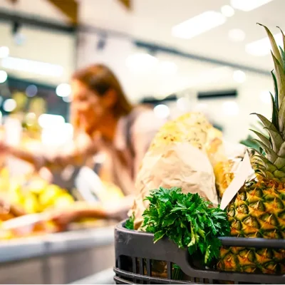 Carrinho cheio de frutas e verduras em supermercado com cliente ao fundo escolhendo produtos, ilustrando a comunicação visual para aumentar ticket médio no supermercado.