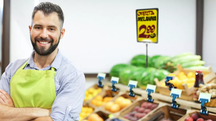 Comerciante sorridente de avental verde em frente a bancada de frutas com placa de oferta visível destacando o preço da melancia.