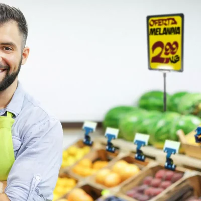 Comerciante sorridente de avental verde em frente a bancada de frutas com placa de oferta visível destacando o preço da melancia.
