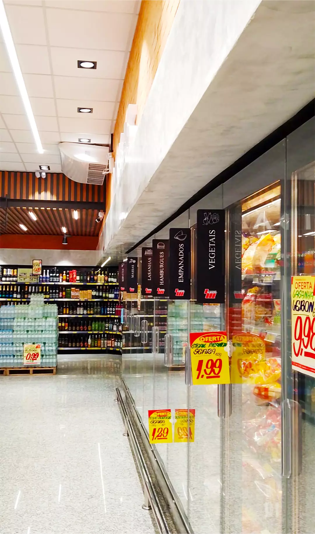 Corredor de supermercado com sinalização suspensa indicando categorias como vegetais, empanados e hambúrgueres, além de etiquetas promocionais em destaque nos freezers.