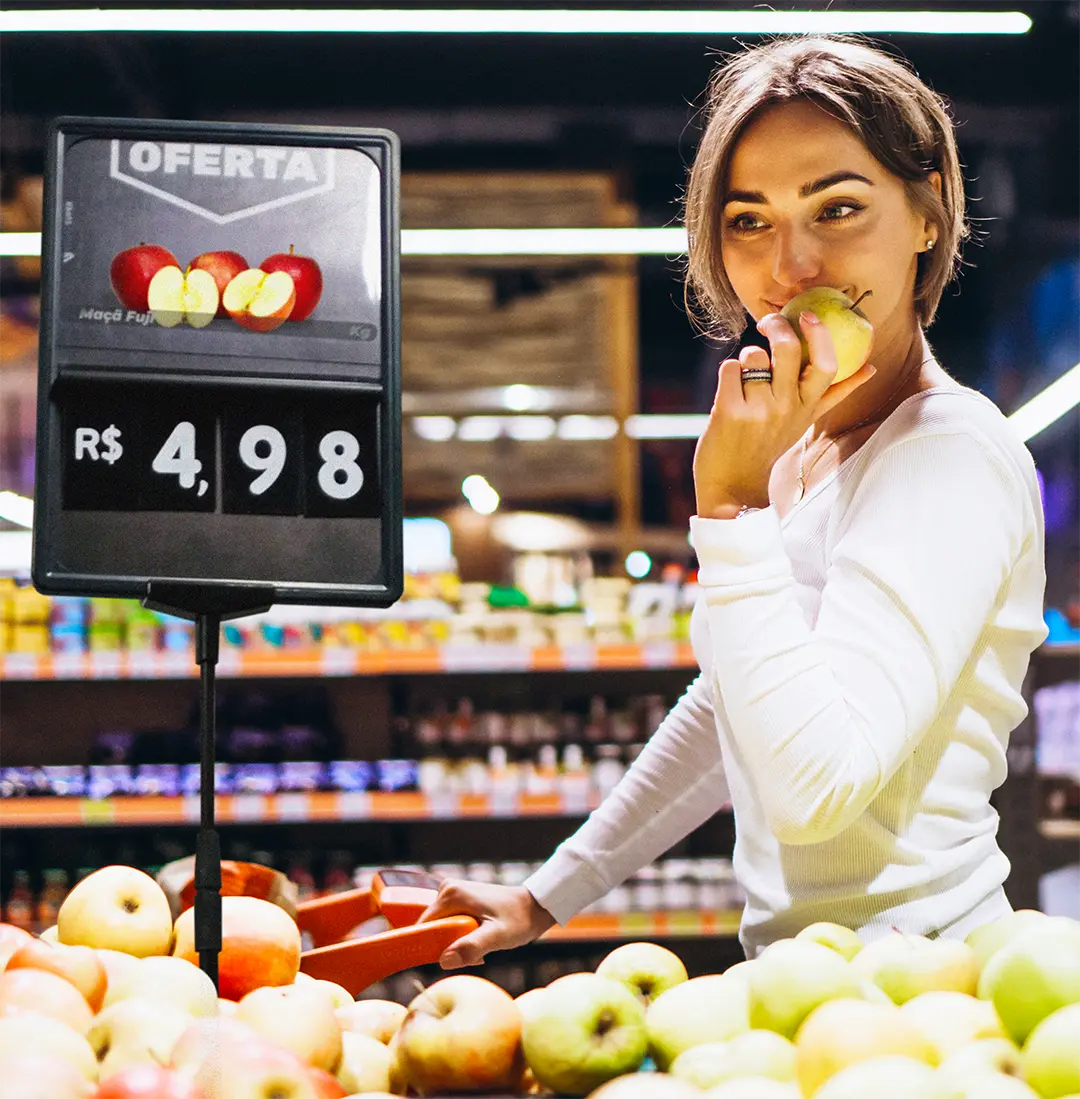Mulher escolhendo maçãs em supermercado ao lado de placa de preço, simbolizando a importância da precificação clara no varejo e da transparência nas informações ao consumidor.