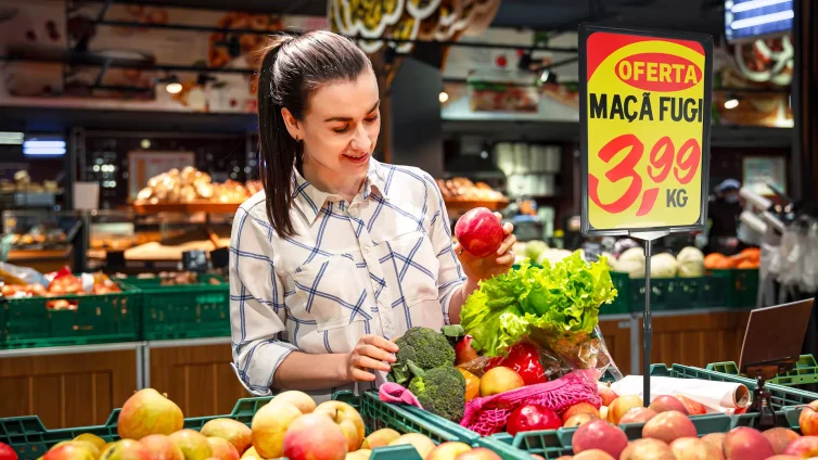 Mulher escolhendo frutas em supermercado com cartaz de oferta destacando preço, representando o uso da comunicação visual no varejo alimentar para aumentar as vendas.