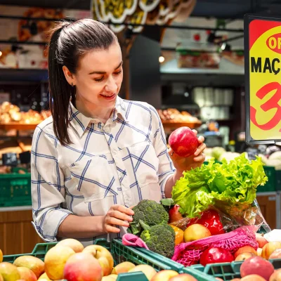 Mulher escolhendo frutas em supermercado com cartaz de oferta destacando preço, representando o uso da comunicação visual no varejo alimentar para aumentar as vendas.