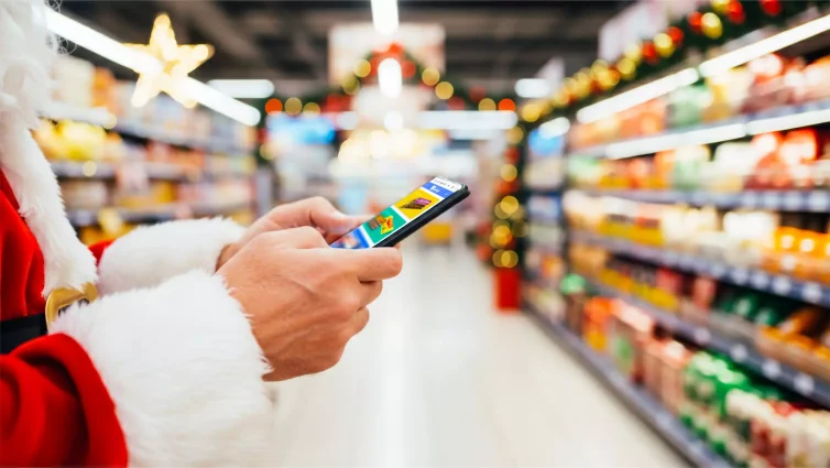Tendências de consumo para o natal. Pessoa vestida com traje de Papai Noel usando o celular enquanto caminha por um supermercado decorado para o Natal.