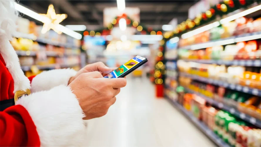 Tendências de consumo para o natal. Pessoa vestida com traje de Papai Noel usando o celular enquanto caminha por um supermercado decorado para o Natal.