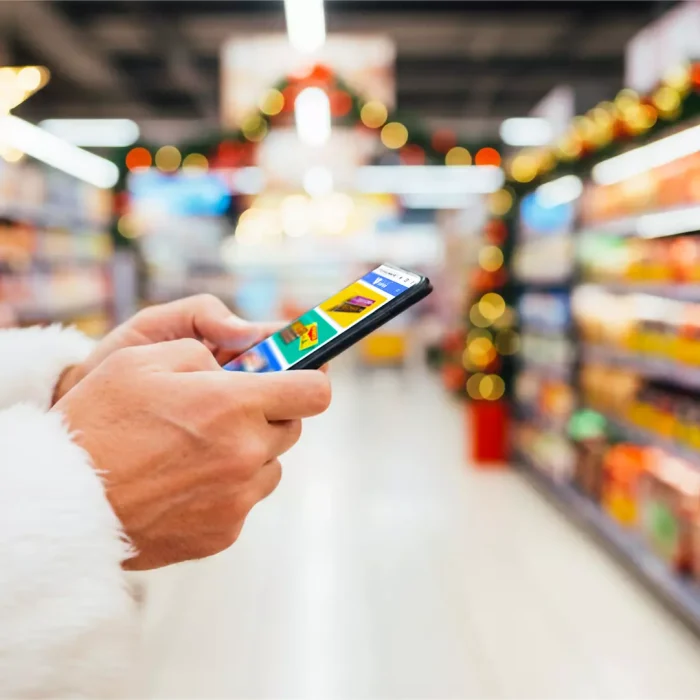 Tendências de consumo para o natal. Pessoa vestida com traje de Papai Noel usando o celular enquanto caminha por um supermercado decorado para o Natal.