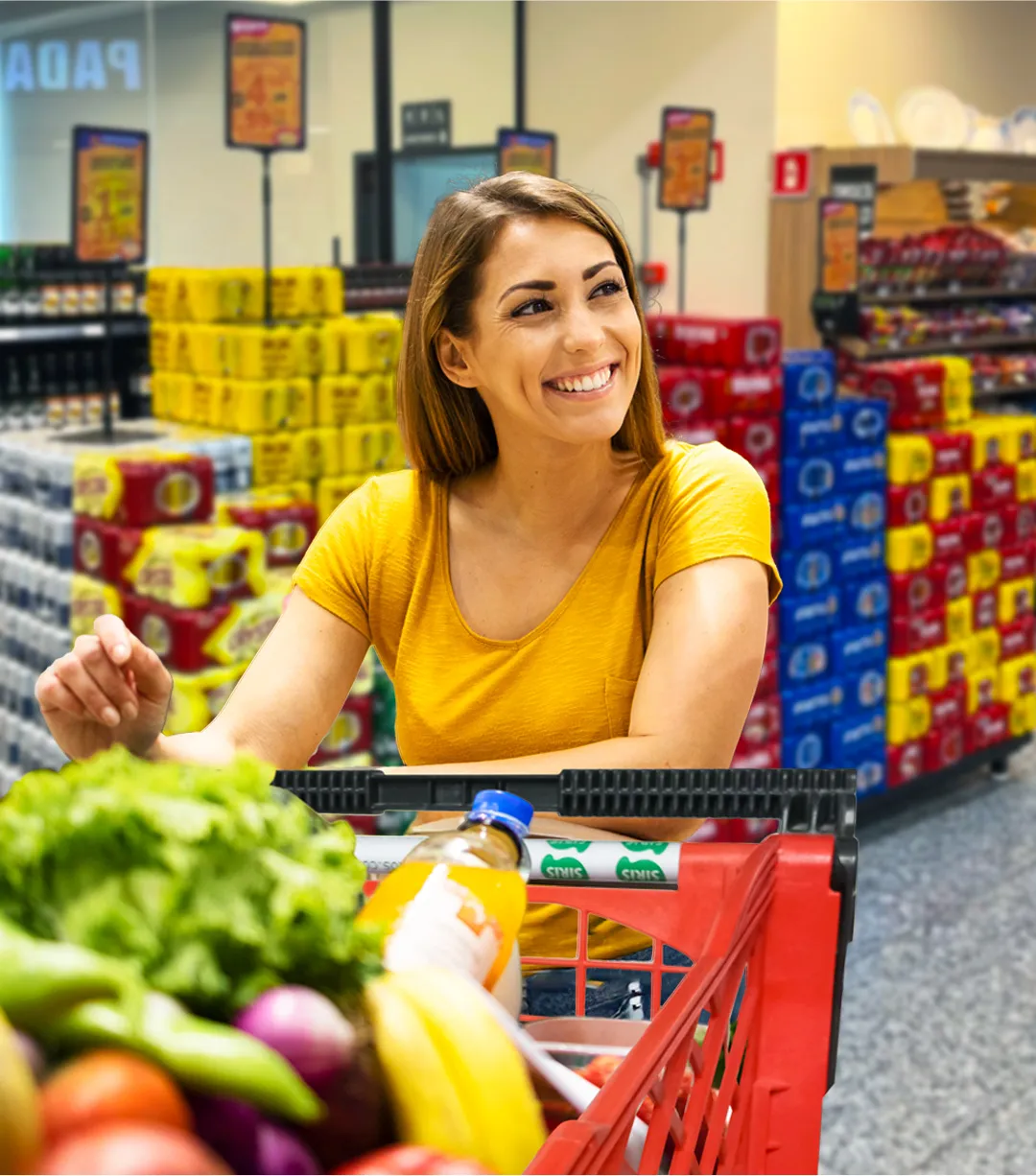 Consumidora sorrindo enquanto faz compras em supermercado com gôndolas organizadas e comunicação visual clara, simbolizando experiência de compra positiva.