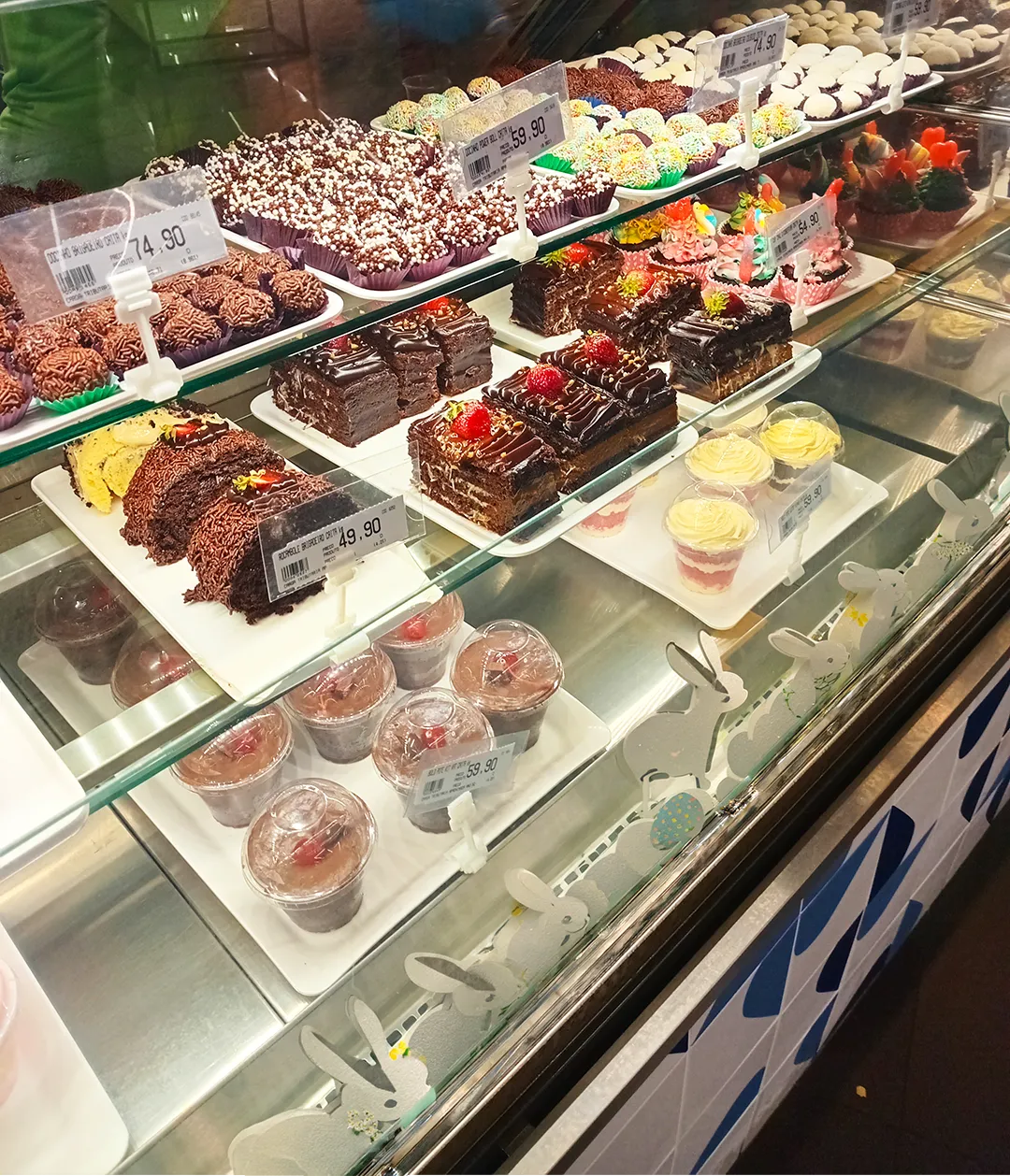 Vitrine de confeitaria com decoração de coelhinhos de Páscoa, exibindo fatias de bolo de chocolate com morango, brigadeiros diversos e sobremesas individuais em potes plásticos.