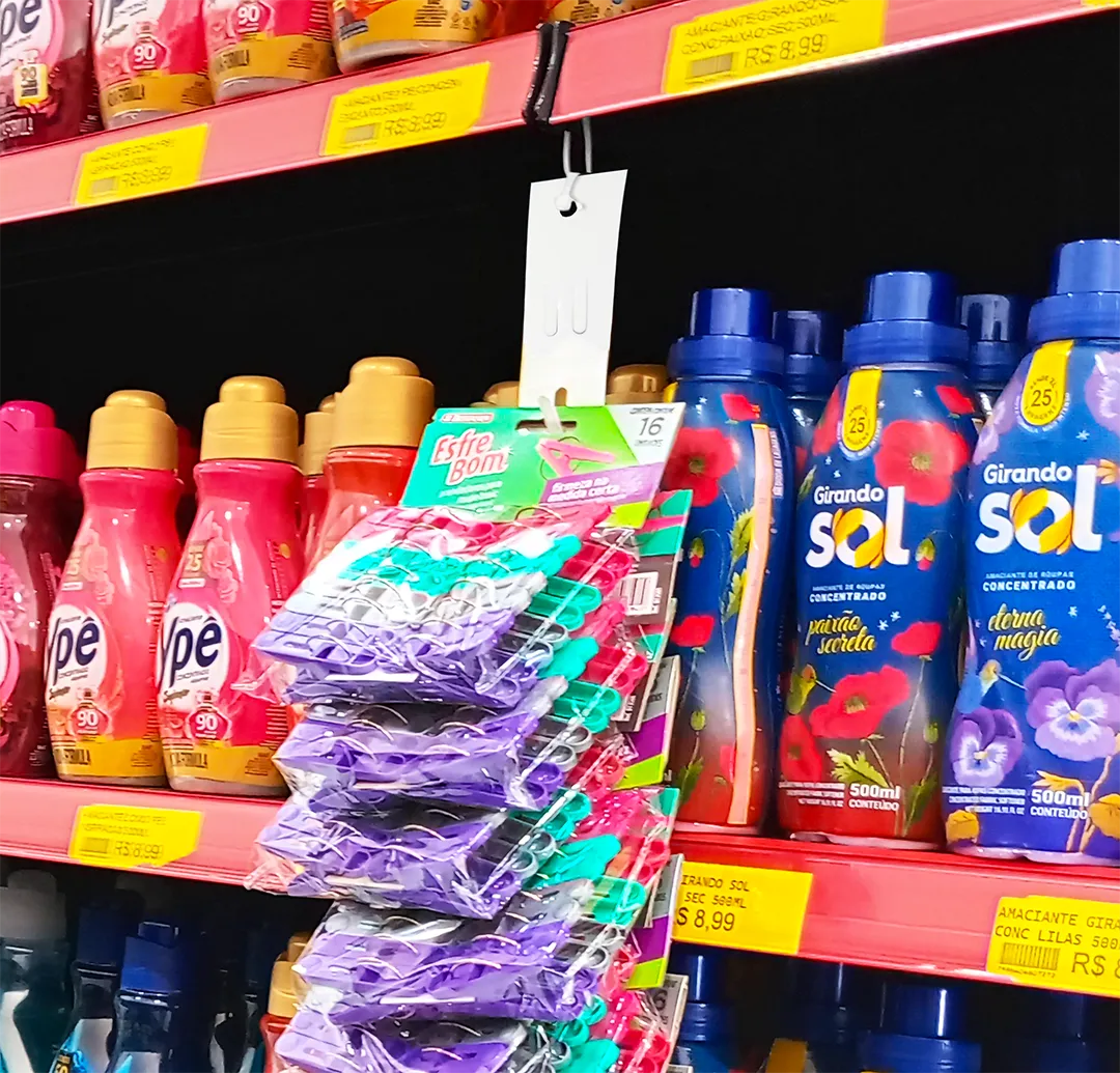 Prateleira de supermercado com diversos frascos de amaciante de roupas das marcas Ypê e Girando Sol. No centro, uma fita "clip strip" pendura pacotes de pregadores de roupa coloridos da marca EsfreBom à frente dos frascos, exemplificando técnica de venda casada.